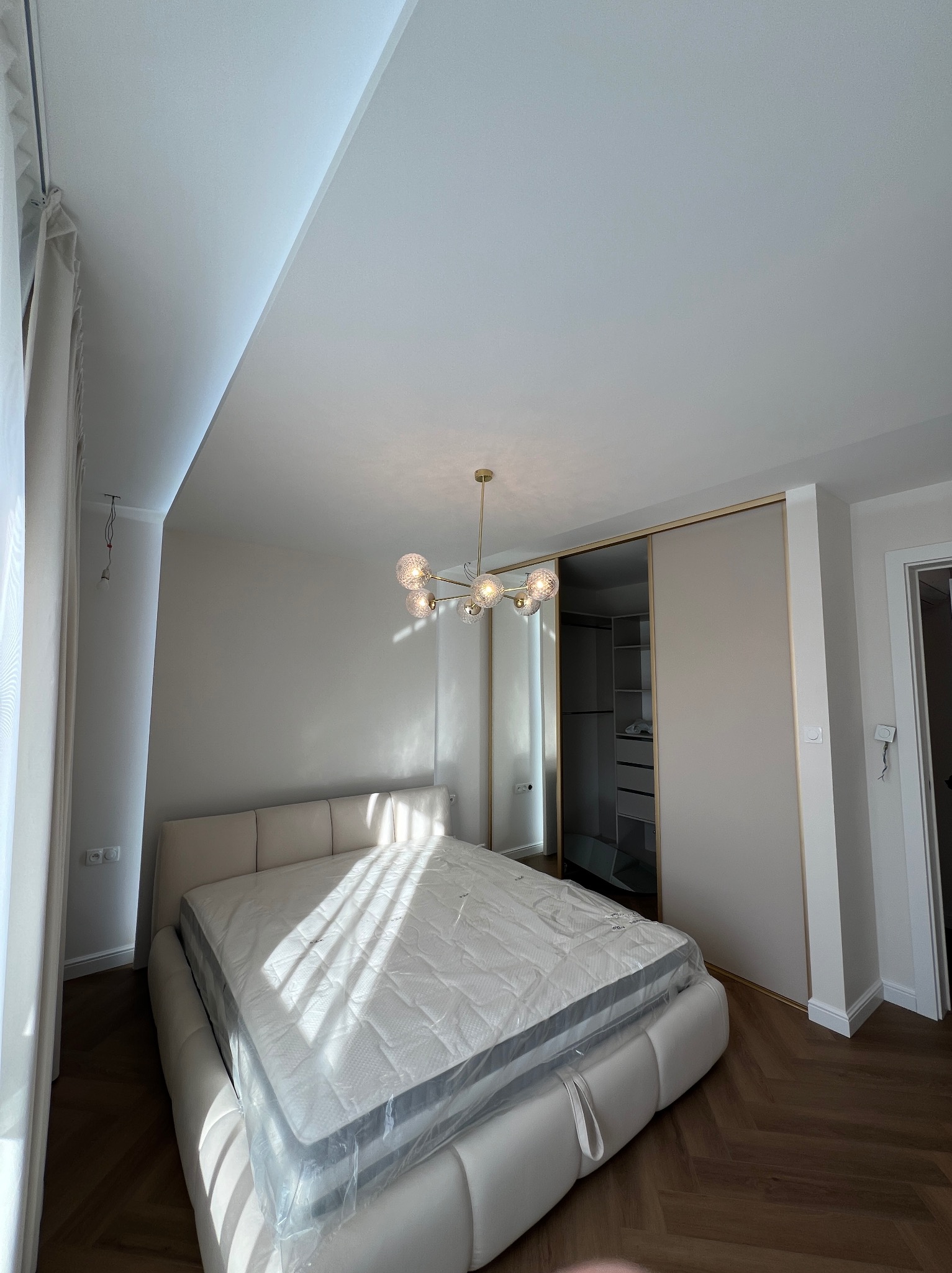Jasna sypialnia z parkietem w jodełkę, łóżkiem z materacem w folii, przesuwną szafą wnękową z lustrem i nowoczesną lampą sufitową.