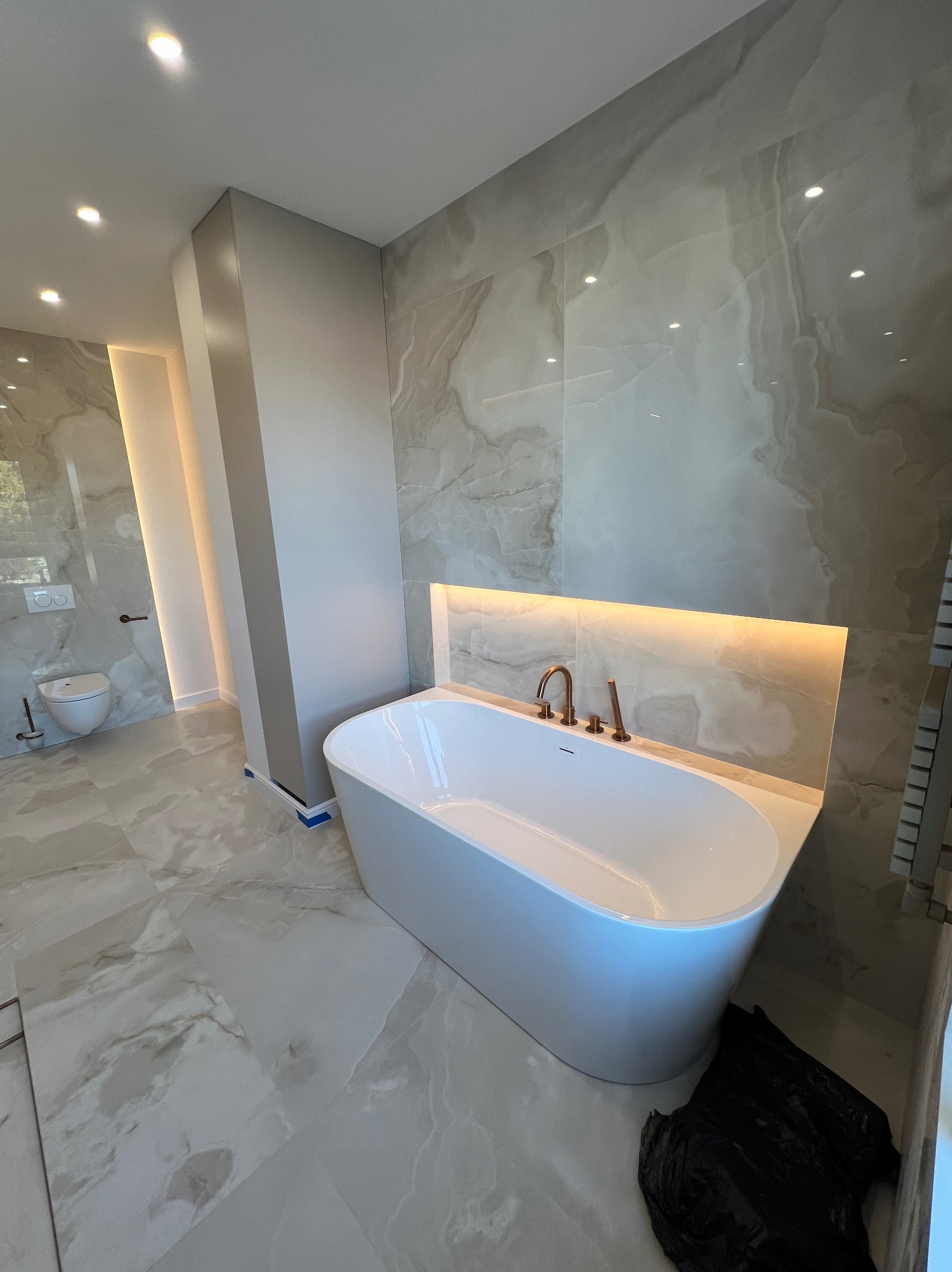 Nowoczesna łazienka z wolnostojącą białą wanną, podświetleniem LED za wanną, marmurowymi płytkami na ścianach i podłodze oraz złotą armaturą, widok na toaletę w tle.