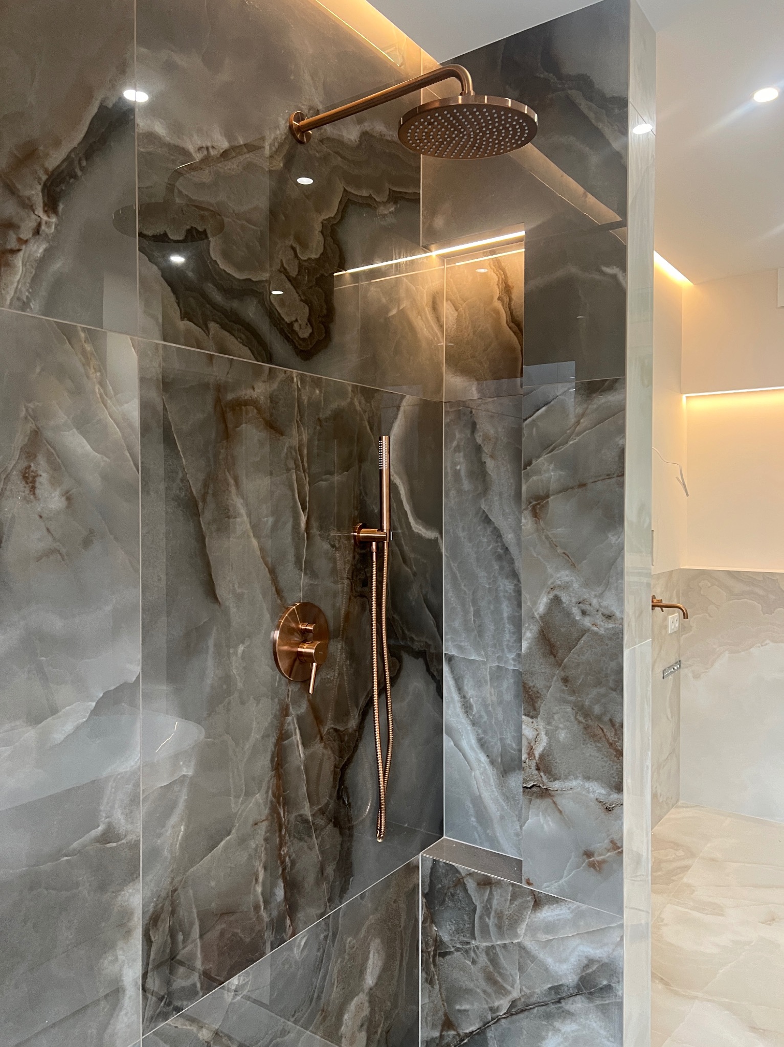 Nowoczesna kabina prysznicowa z marmurowymi płytkami w odcieniach szarości i brązu, armatura w kolorze różowego złota, oświetlenie LED wzdłuż sufitu i ściany.