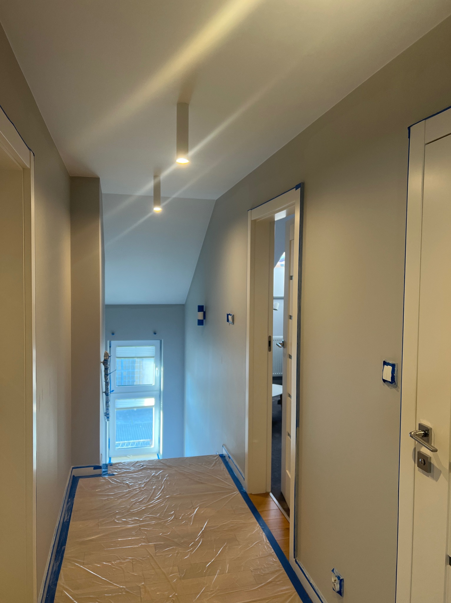 Korytarz w trakcie malowania: ściany w szarym odcieniu zabezpieczone taśmą malarską, podłoga przykryta folią ochronną, widoczne lampy sufitowe.