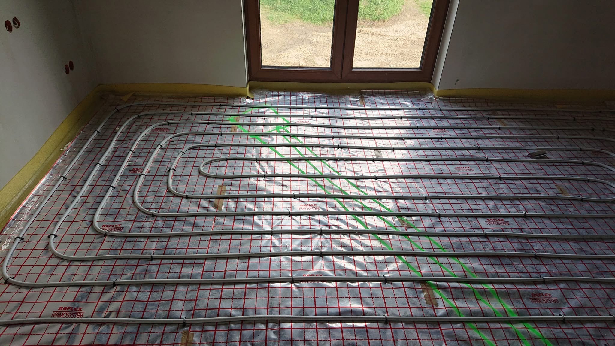Układanie ogrzewania podłogowego na folii z siatką, widoczne rurki i zielone linie wyznaczające trasy, żółta taśma przy ścianie, okno w tle.