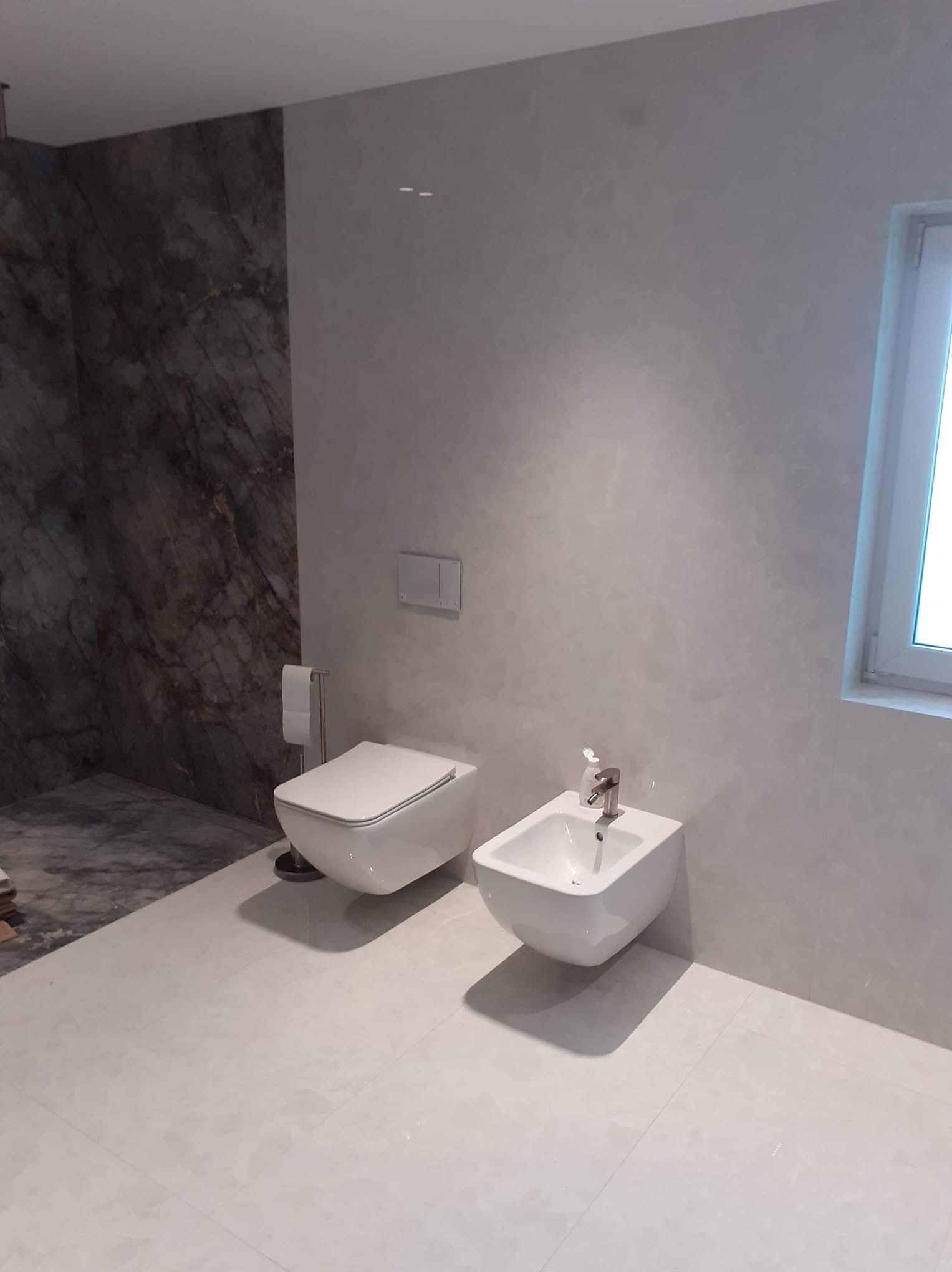 Nowoczesna łazienka z marmurową ścianą, podwieszaną toaletą i bidetem na jasnych płytkach podłogowych, minimalistyczny design.