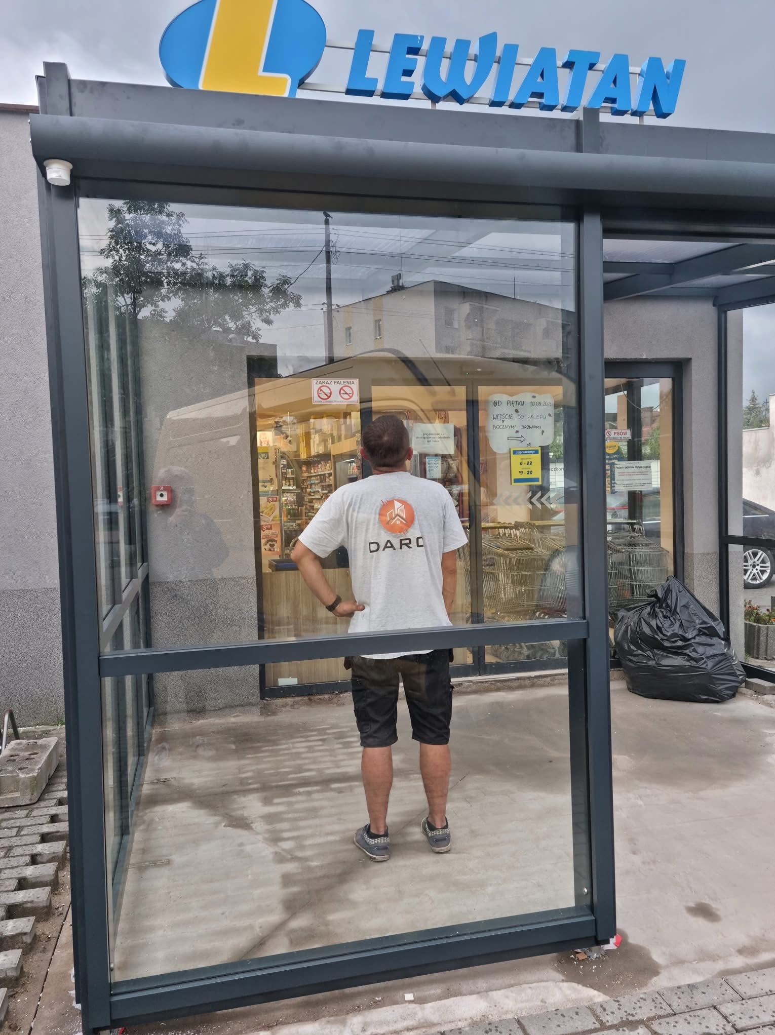 Zewnętrzna szklana konstrukcja przy wejściu do sklepu Lewiatan, widok od tyłu na mężczyznę w koszulce z logo DARD, odblask na szybie, czarny worek na śmieci, wózki sklepowe w tle.