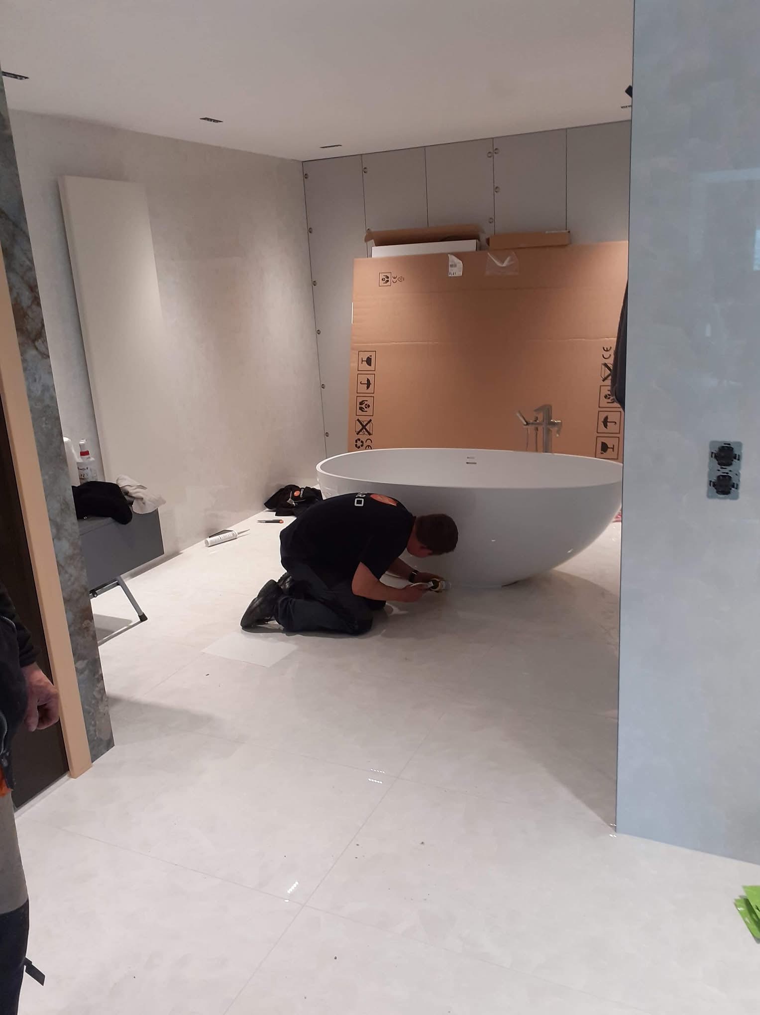 Pracownik na kolanach silikonuje białą, wolnostojącą wannę w minimalistycznej łazience z szarymi płytkami i kartonami w tle.