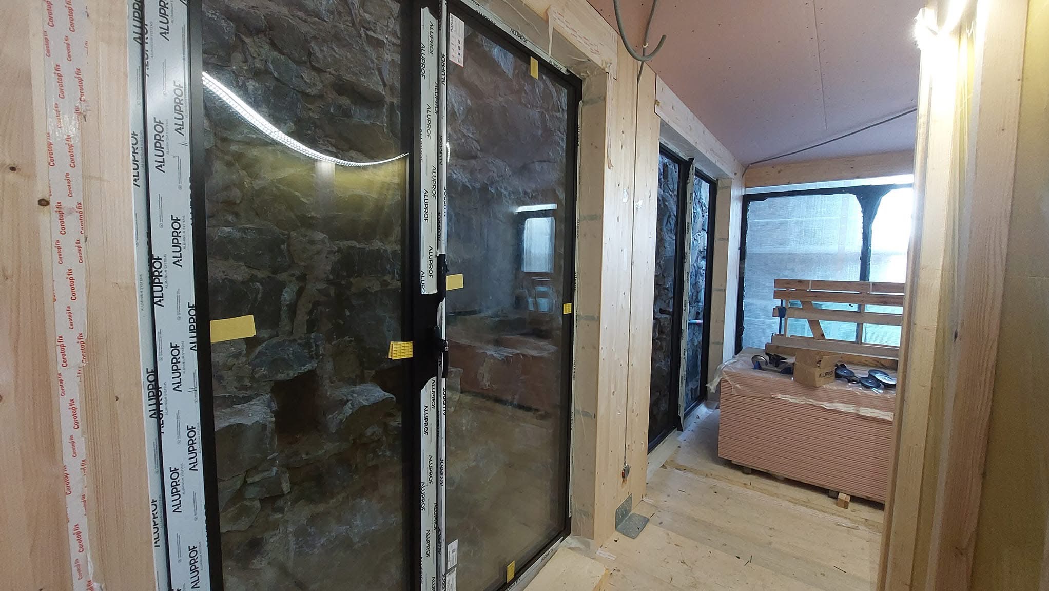 Montaż okien z czarnymi ramami Alupof w surowym wnętrzu z kamienną ścianą, widoczne deski i materiały budowlane, etap wykończenia.