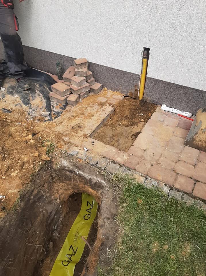 Wykop w ziemi z widoczną żółtą taśmą ostrzegawczą 'GAZ' obok fragmentu chodnika z kostki, przy ścianie budynku z wystającą rurą gazową.