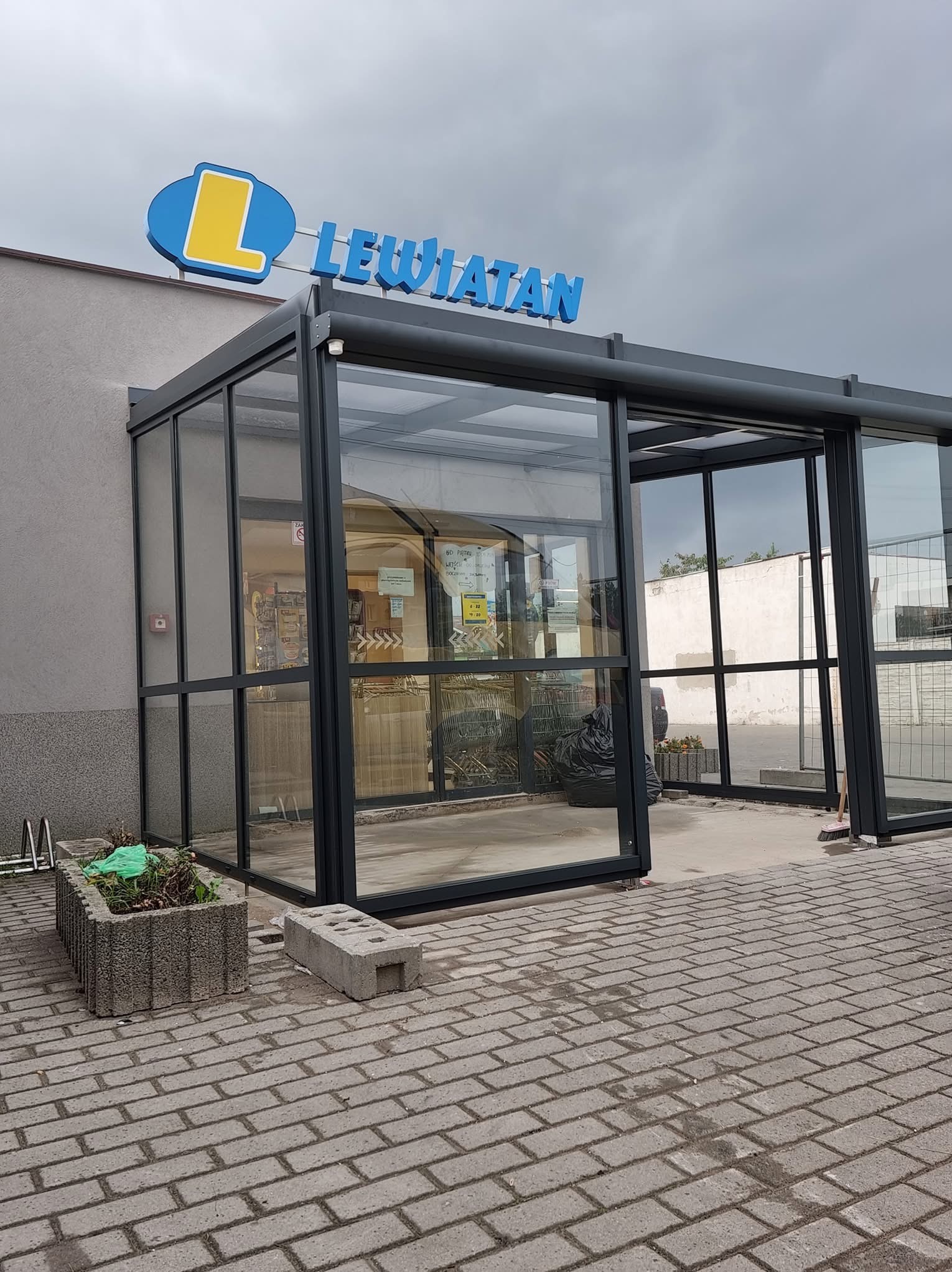 Zewnętrzny widok przeszklonego przedsionka wejścia do sklepu Lewiatan z widocznym logo i kostką brukową na pierwszym planie.