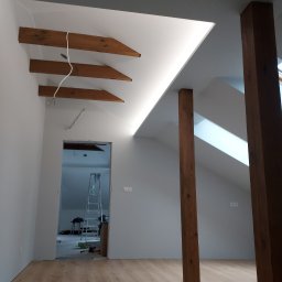 Mateusz Rypień - Poddasze po remoncie z drewnianymi belkami stropowymi, oświetleniem LED w szczycie dachu, drewnianymi słupami i panelami podłogowymi. W tle widoczna drabina i narzędzia.