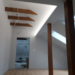 Poddasze po remoncie z drewnianymi belkami stropowymi, oświetleniem LED w szczycie dachu, drewnianymi słupami i panelami podłogowymi. W tle widoczna drabina i narzędzia.