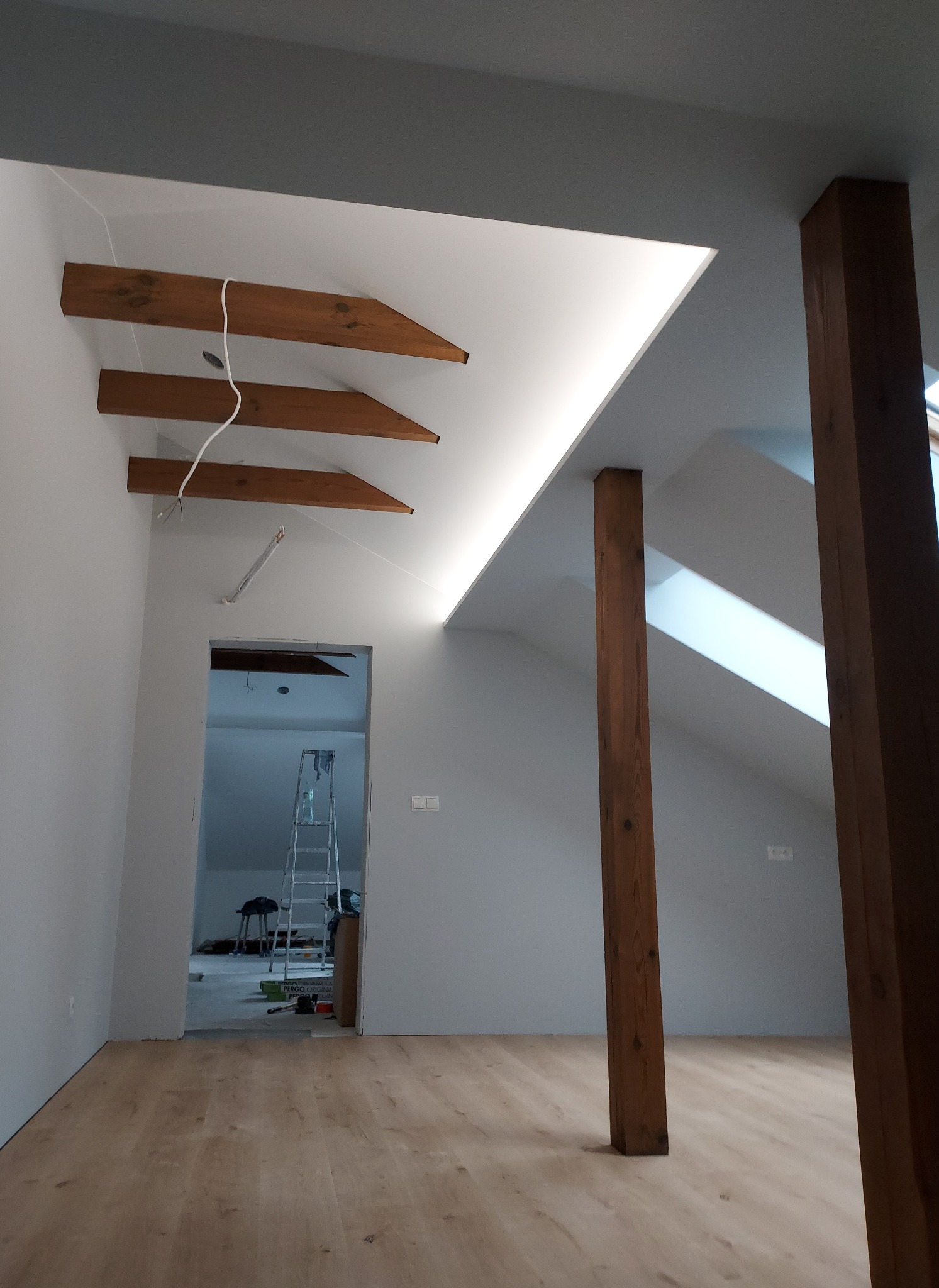 Poddasze po remoncie z drewnianymi belkami stropowymi, oświetleniem LED w szczycie dachu, drewnianymi słupami i panelami podłogowymi. W tle widoczna drabina i narzędzia.