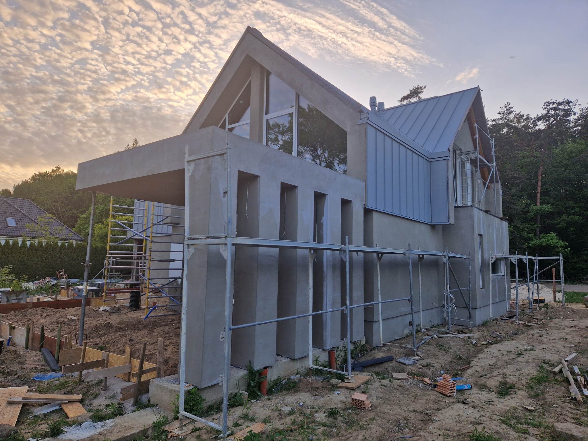 Surowy stan domu jednorodzinnego z szarą blachą na dachu, widoczne rusztowania i przygotowany teren pod fundamenty, niebo z obłokami o zachodzie słońca