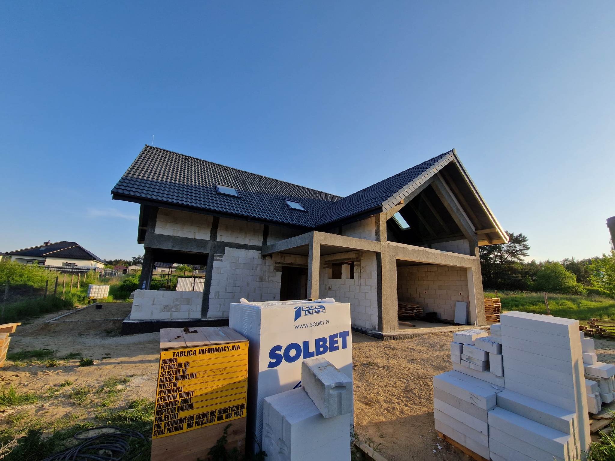 Surowy stan domu jednorodzinnego z bloczków betonu komórkowego Solbet, widoczna tablica informacyjna budowy z danymi inwestycji.