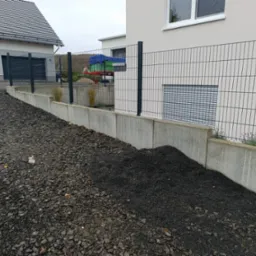 Nowoczesne ogrodzenie panelowe w kolorze grafitowym, zamontowane na betonowej podmurówce, z widocznymi elementami posesji w tle i nasypanym kamieniem ozdobnym u podstawy.