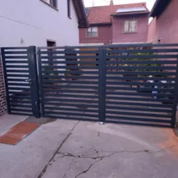 Nowoczesna, pozioma brama metalowa z furtką, pomalowana na ciemnoszary kolor, zamontowana przy ceglanym słupku i betonowym podjeździe.