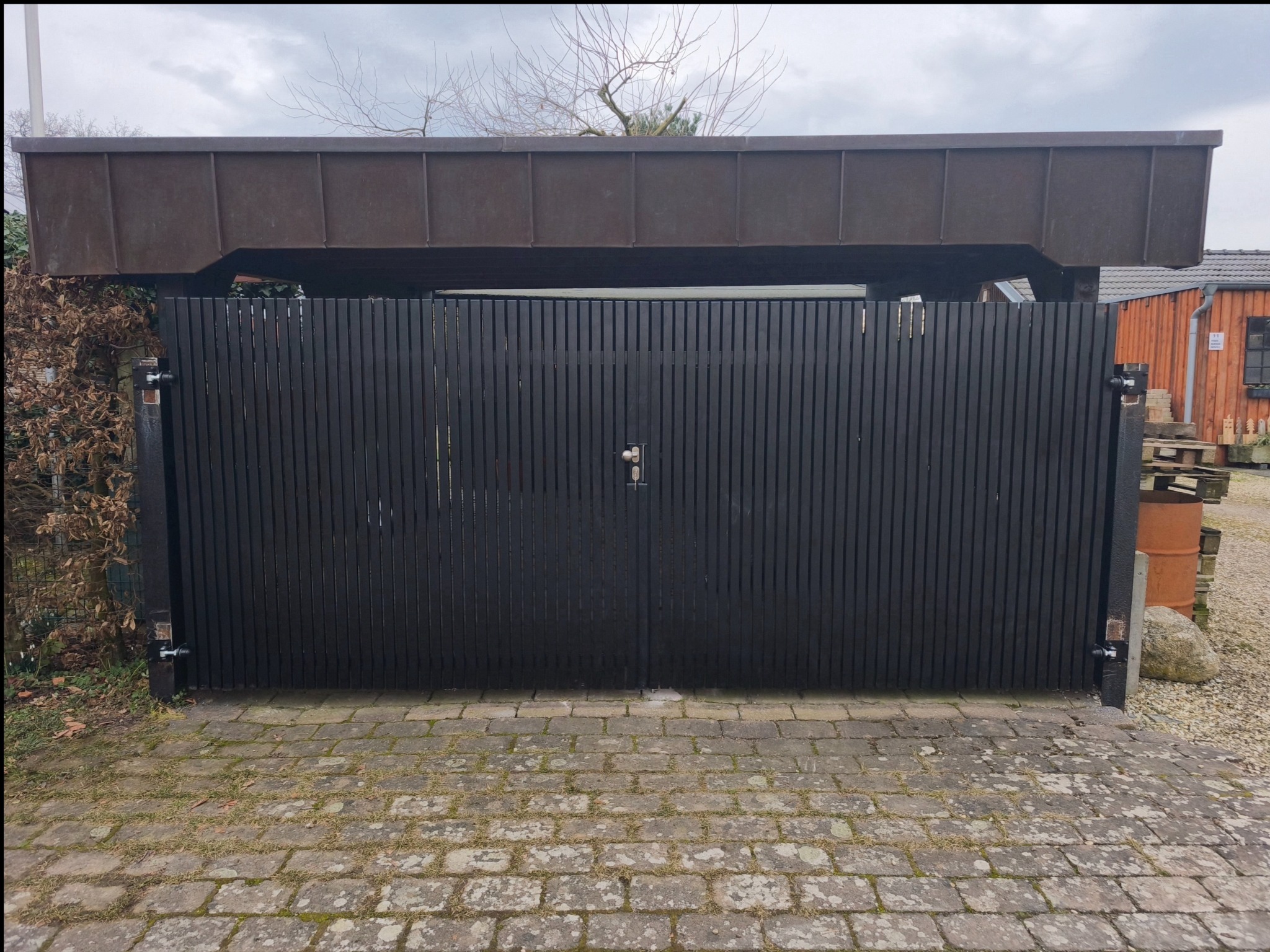 Nowoczesna, czarna, dwuskrzydłowa brama wjazdowa z pionowych desek, zadaszona konstrukcją z blachy, na tle kostki brukowej.
