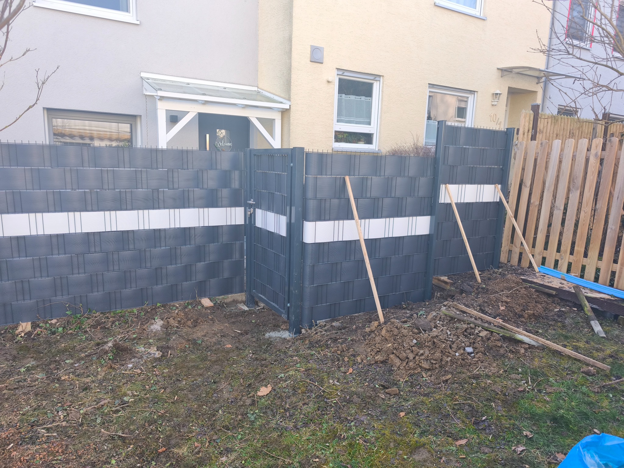 W trakcie montażu szarego, panelowego ogrodzenia z białą linią, widoczne drewniane podpory, ziemia i fragment drewnianego płotu sąsiada.