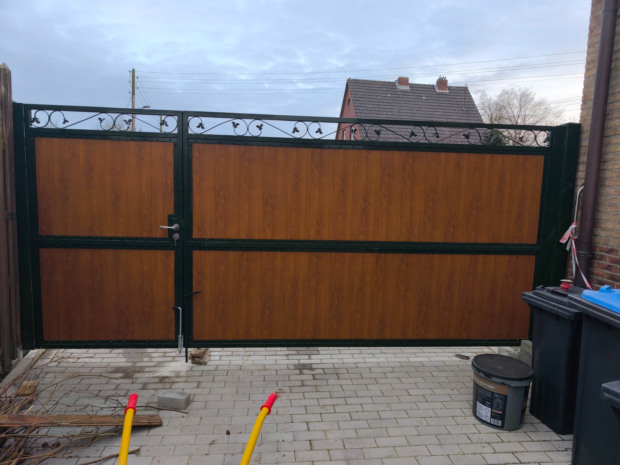 Dwuskrzydłowa brama wjazdowa z drewnopodobnymi panelami i metalową ramą z ozdobnymi elementami, widok z zewnątrz na posesję z kostki brukowej, obok stoją pojemniki na śmieci i wiadro farby.