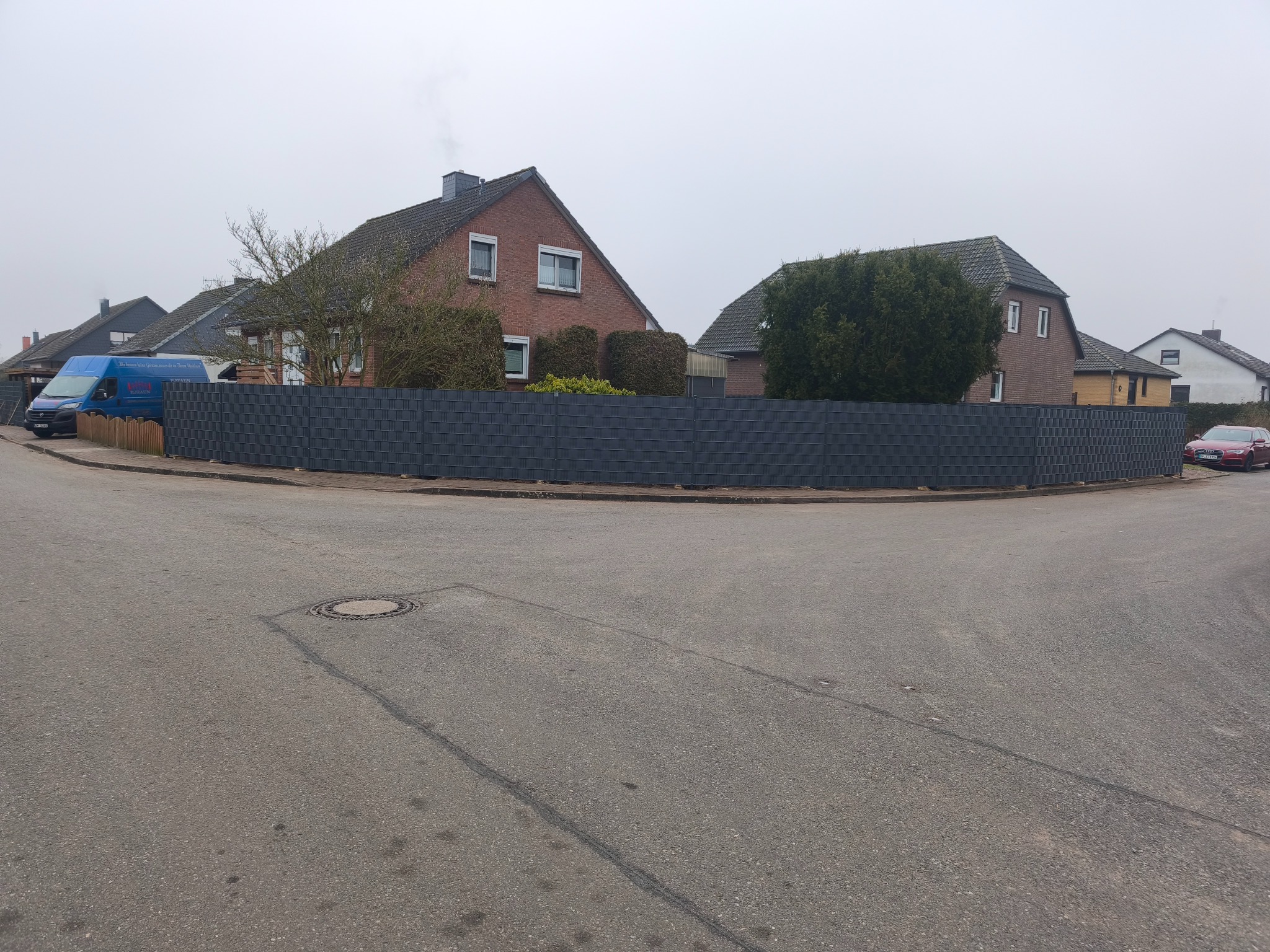 Szary panelowy płot odgradzający posesje od skrzyżowania ulic w pochmurny dzień, widoczny samochód dostawczy i osobowy.