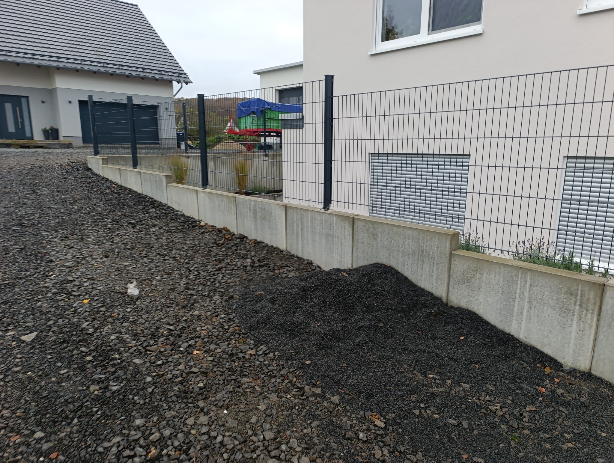 Nowoczesne ogrodzenie panelowe w kolorze grafitowym, zamontowane na betonowej podmurówce, z widocznymi elementami posesji w tle i nasypanym kamieniem ozdobnym u podstawy.