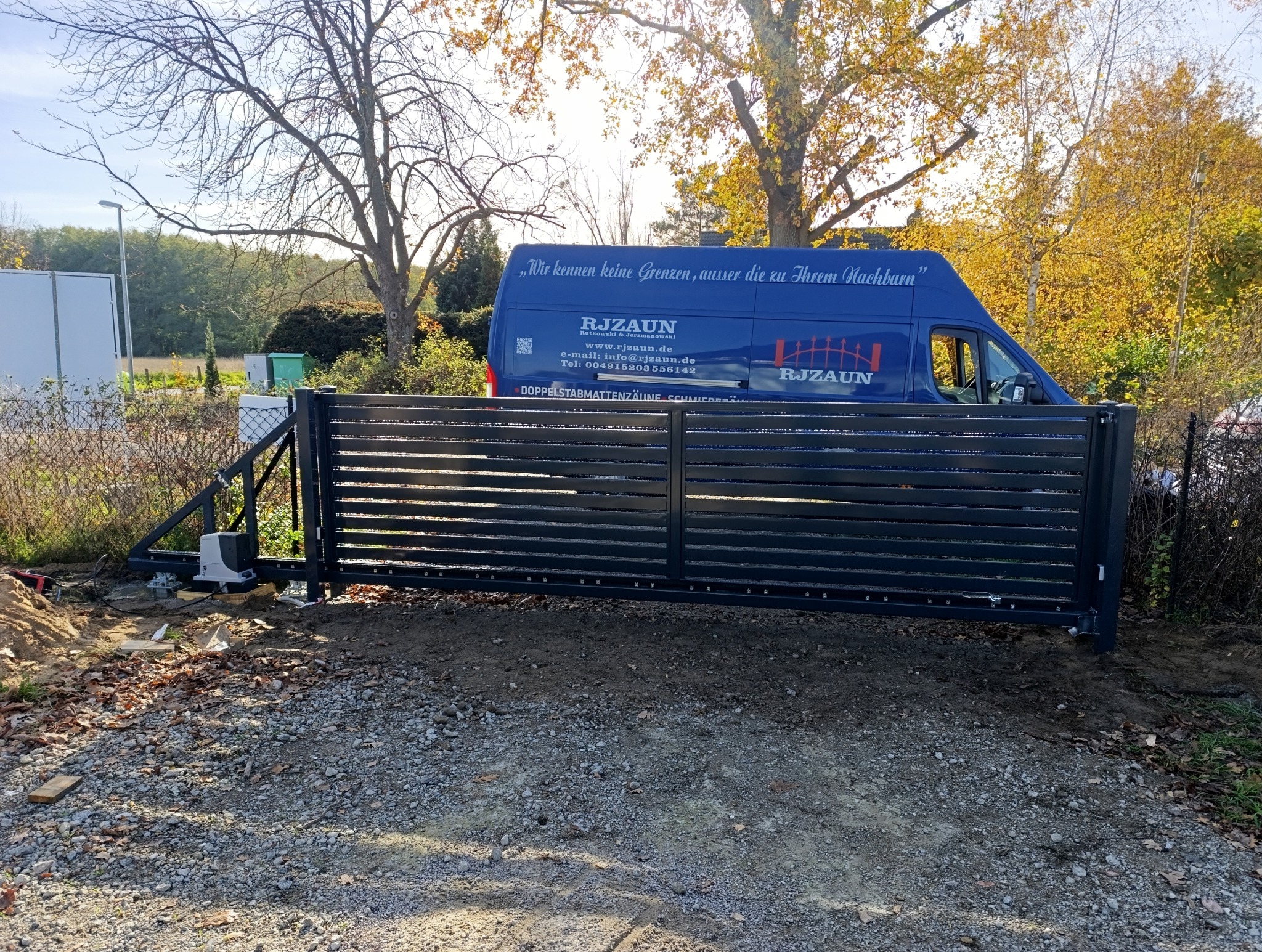 Automatyczna brama przesuwna z poziomych profili metalowych, widoczny napęd i częściowo odsłonięty fundament, w tle firmowy bus z logo i danymi kontaktowymi.