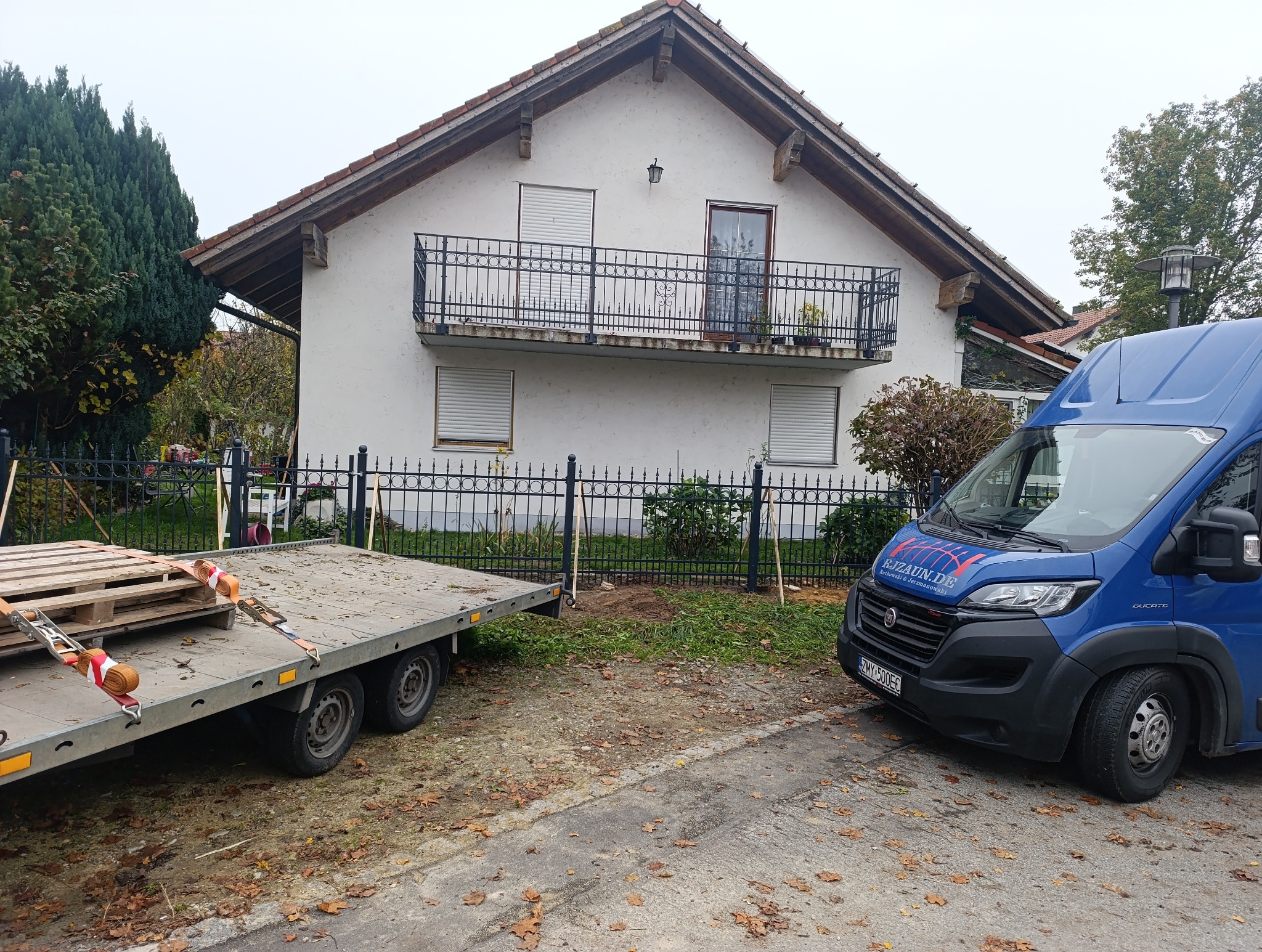 Przygotowania do montażu ogrodzenia metalowego: przyczepa z paletami i słupkami, oraz firmowy bus na tle domu z balkonem.