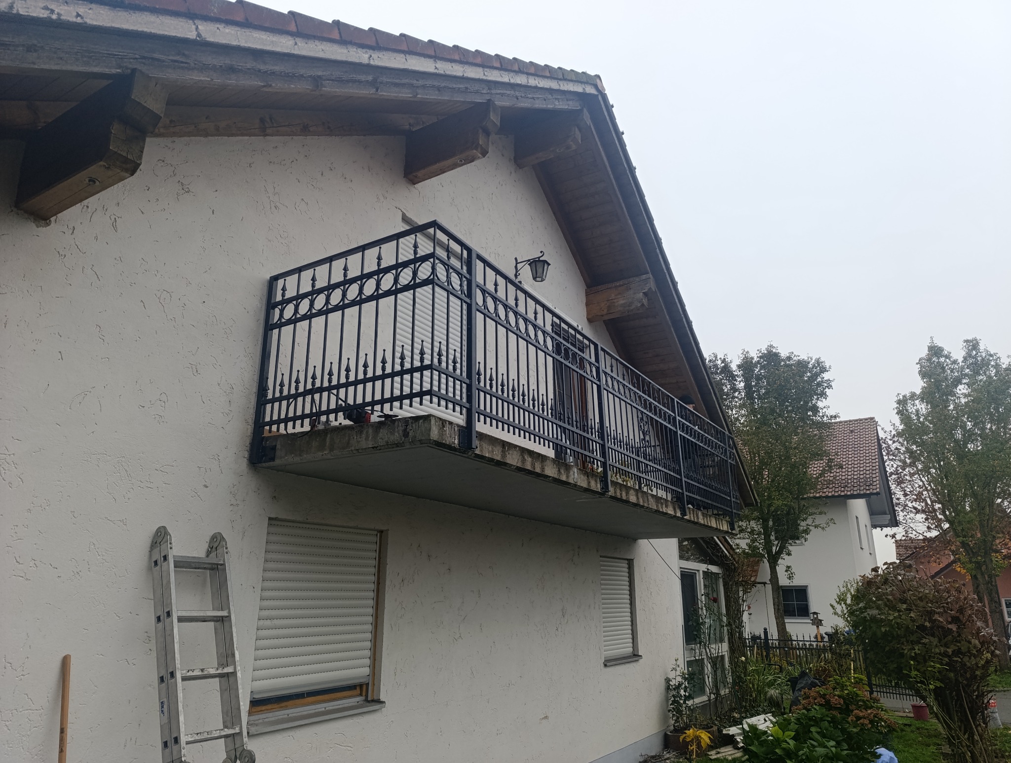 Czarna, metalowa balustrada balkonu z ozdobnymi elementami, przymocowana do ściany budynku z fakturą, drabina oparta o ścianę obok okna z roletą.