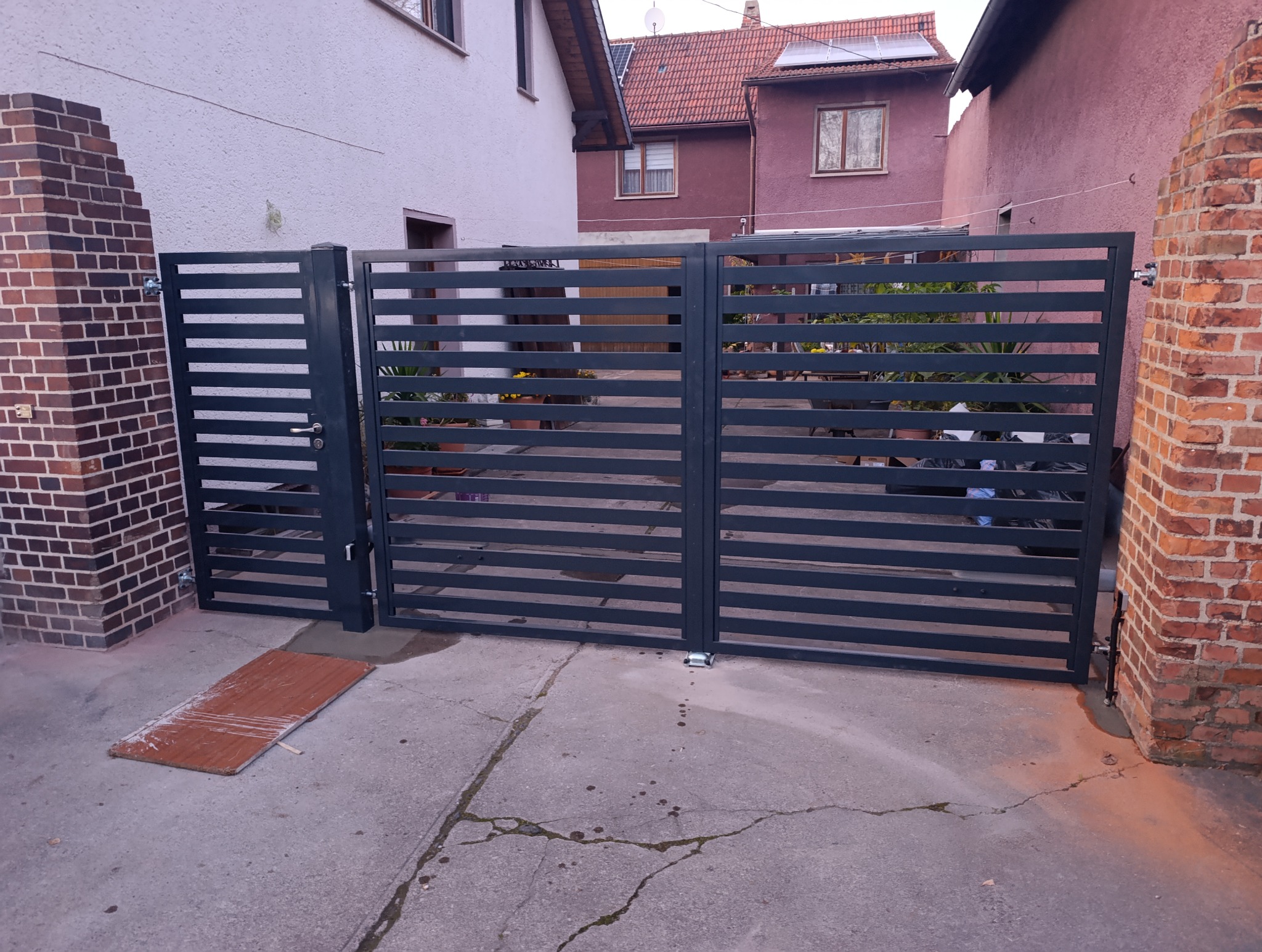 Nowoczesna, pozioma brama metalowa z furtką, pomalowana na ciemnoszary kolor, zamontowana przy ceglanym słupku i betonowym podjeździe.