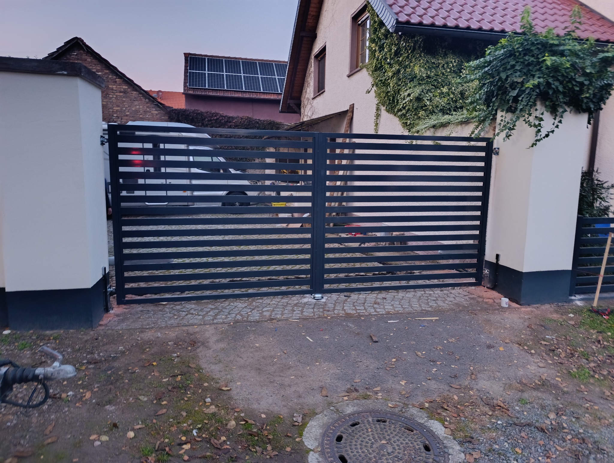 Nowoczesna, pozioma brama metalowa w kolorze grafitowym, zamontowana między dwoma białymi słupkami, widok na posesję z kamperem w tle.