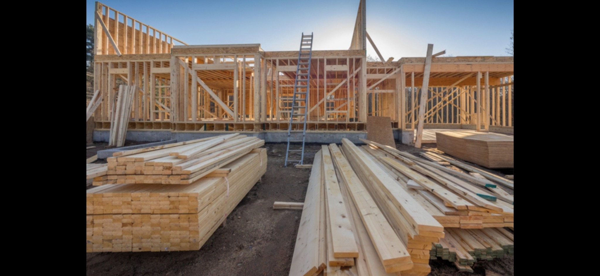 Konstrukcja domu w trakcie budowy z drewnianym szkieletem, widoczne fundamenty i stosy drewna oraz drabina oparta o budynek w słoneczny dzień.