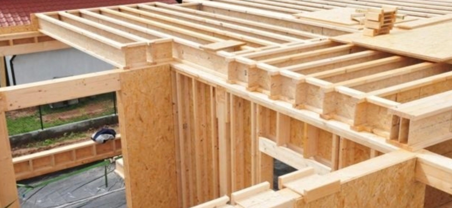 Surowa konstrukcja domu szkieletowego z widocznymi belkami stropowymi i ścianami z płyt OSB, ukazująca detale łączeń i strukturę drewna.