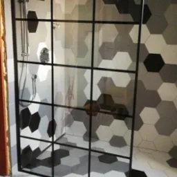 Kabina prysznicowa z czarnymi profilami, heksagonalne płytki w odcieniach szarości na ścianach i podłodze, marmurowe płytki pod kabiną, deszczownica zamontowana na suficie.