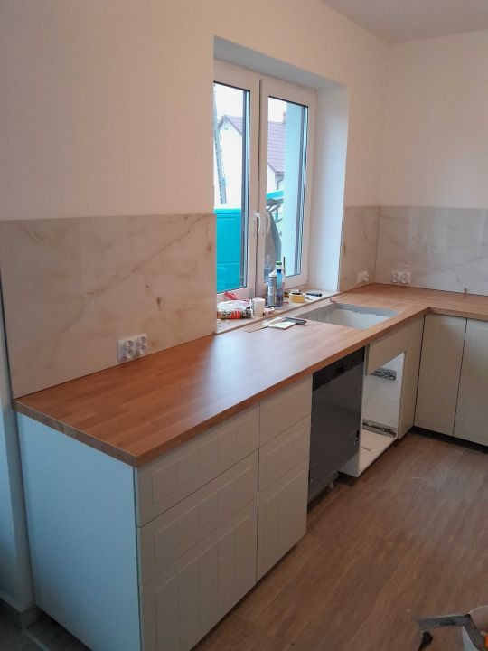 Montaż kuchni: białe szafki z drewnianym blatem, marmurowe panele ścienne, okno z widokiem na domy w tle. Częściowo zamontowana zmywarka.