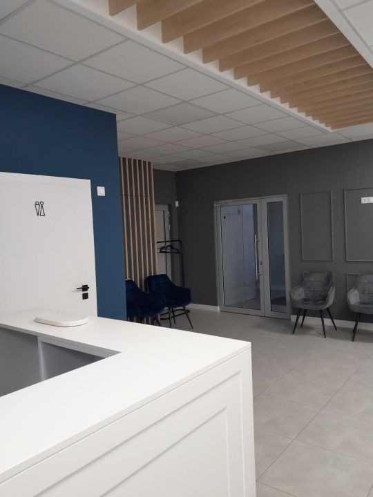 Nowoczesne wnętrze lokalu usługowego w Ulanowie, z białą recepcją, niebieską ścianą z piktogramami toalet, szarą podłogą z płytek i drewnianym elementem dekoracyjnym na suficie.
