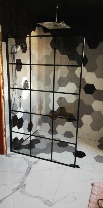 Kabina prysznicowa z czarnymi profilami, heksagonalne płytki w odcieniach szarości na ścianach i podłodze, marmurowe płytki pod kabiną, deszczownica zamontowana na suficie.