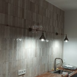 RUSLAN KOVAL MDRinstalelektryka - Rząd czterech stożkowych lamp w stylu industrialnym zamontowanych na rurze nad umywalką w kuchni z ceramiczną ścianą w odcieniach beżu i szarości.