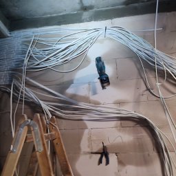 RUSLAN KOVAL MDRinstalelektryka - Rozprowadzenie kabli elektrycznych po ścianach i suficie surowego pomieszczenia, widoczna drabina, wiertarka i kombinerki w ścianie.