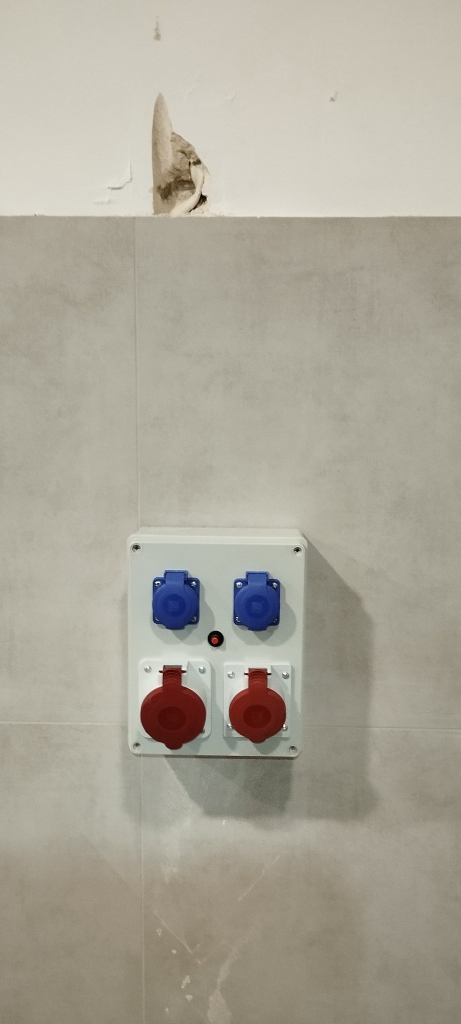 Natynkowa skrzynka elektryczna z gniazdami siłowymi (czerwone i niebieskie) zamontowana na wykafelkowanej ścianie, widoczne uszkodzenie tynku powyżej.