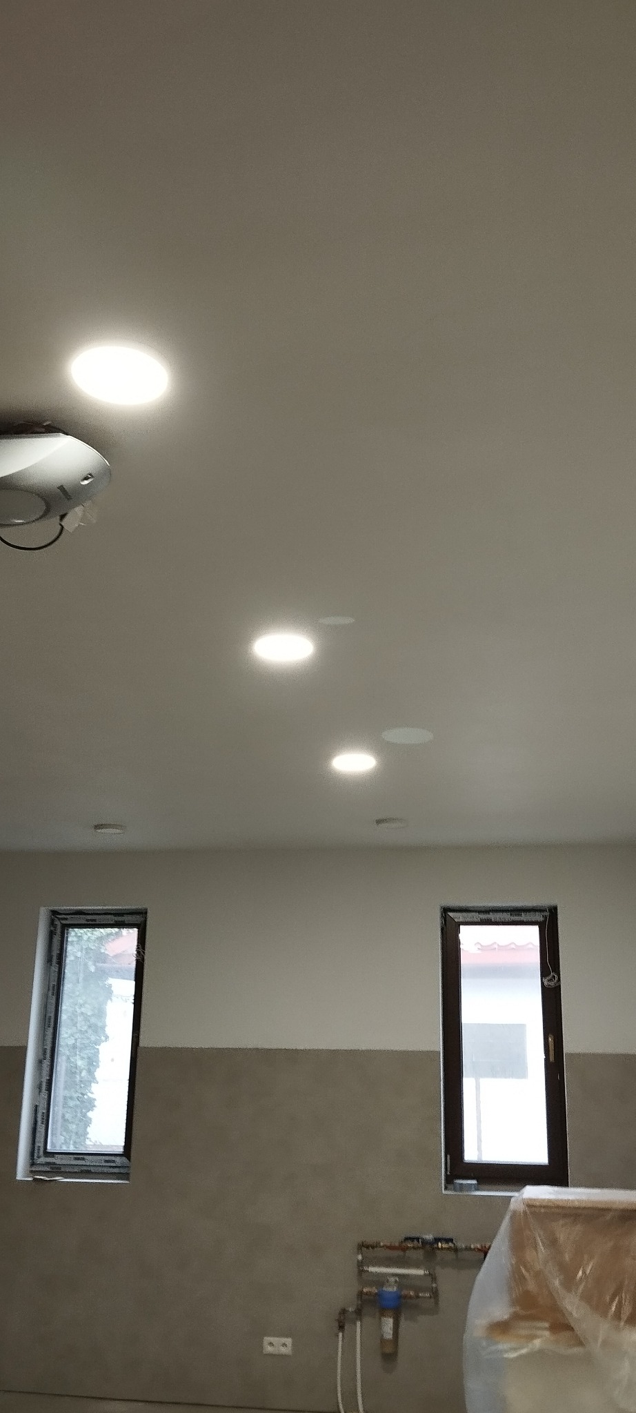 Wnętrze w trakcie wykańczania: biały sufit z zamontowanymi okrągłymi lampami LED, okna z widokiem na zieleń, fragment instalacji wodnej przy ścianie.
