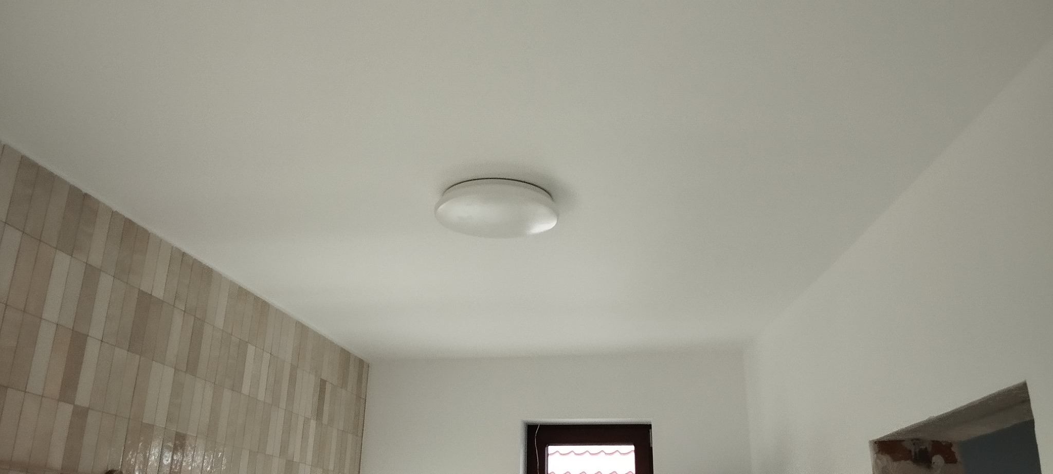 Okrągła lampa sufitowa zamontowana na białym suficie w pomieszczeniu z beżowymi płytkami na ścianie i widocznym fragmentem okna.