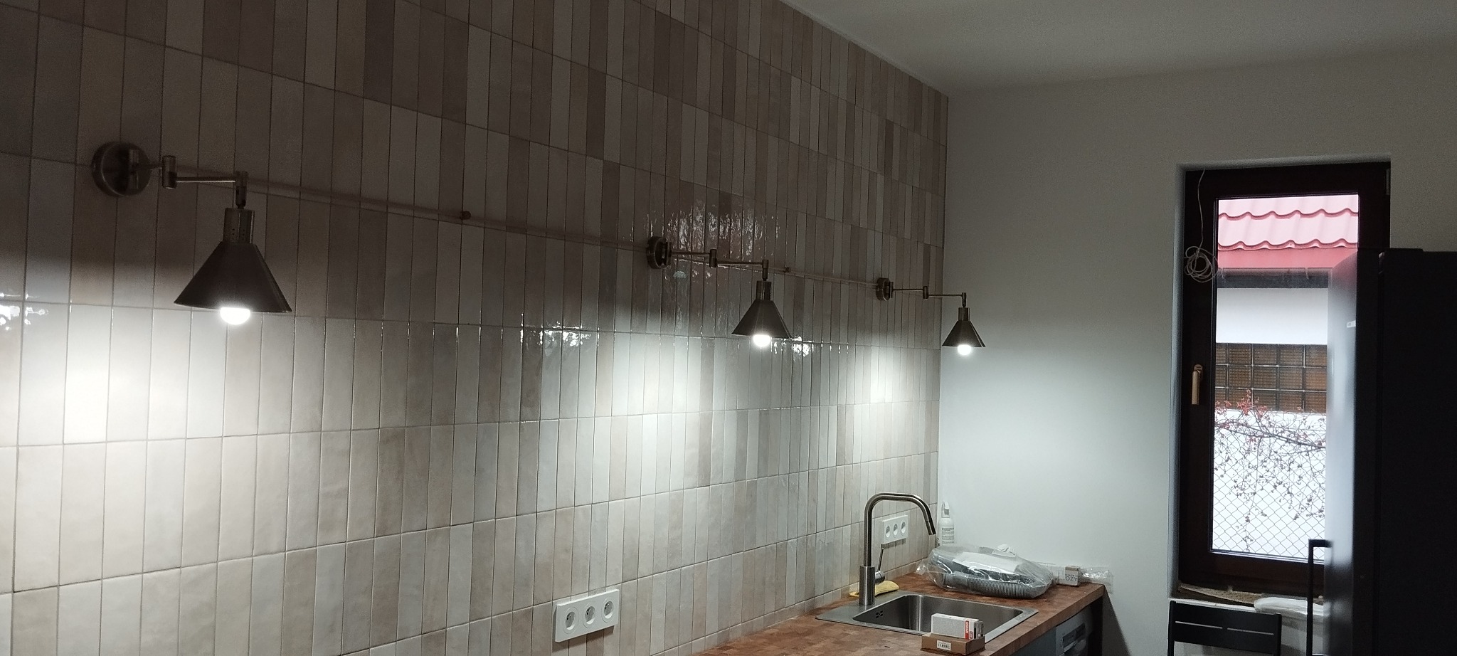 Rząd czterech stożkowych lamp w stylu industrialnym zamontowanych na rurze nad umywalką w kuchni z ceramiczną ścianą w odcieniach beżu i szarości.