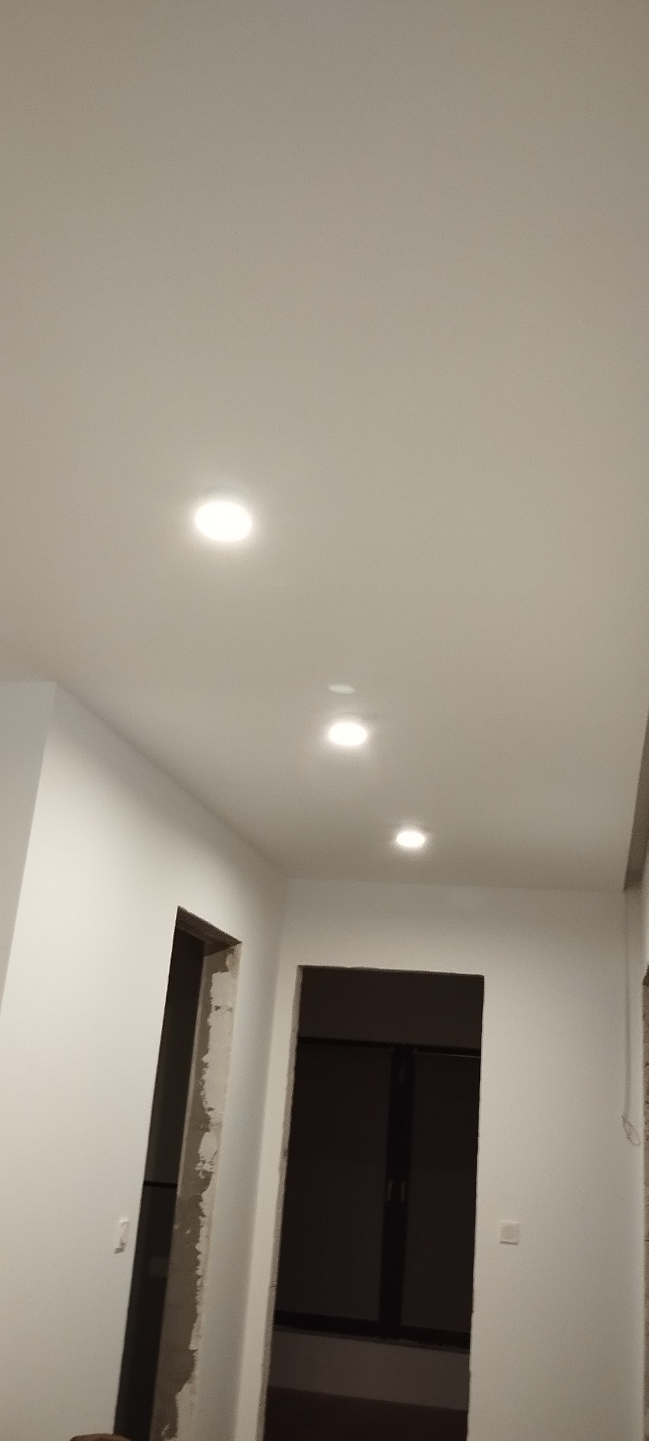 Rząd trzech okrągłych lamp LED wpuszczonych w biały sufit, widok z korytarza w trakcie wykańczania mieszkania w Warszawie, z widocznymi otworami drzwiowymi.
