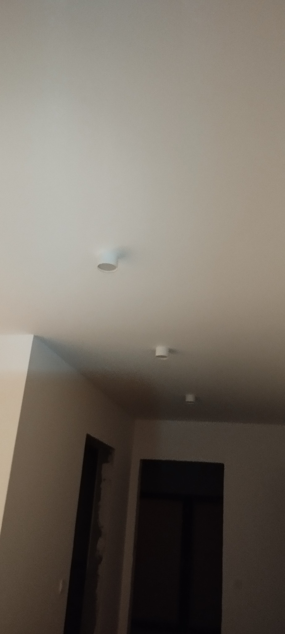 Biały sufit z zamontowanymi trzema okrągłymi lampami sufitowymi typu downlight w nowym budynku, widoczne fragmenty ścian i otwory drzwiowe w stanie surowym.