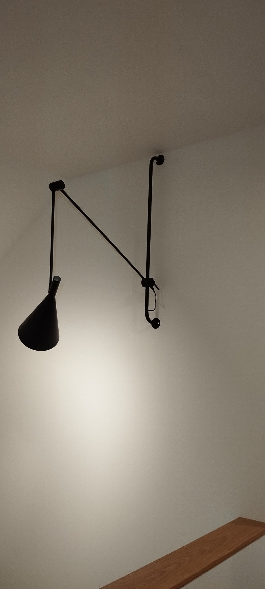 Czarna, regulowana lampa ścienna w stylu industrialnym, oświetlająca fragment białej ściany i drewnianego blatu.