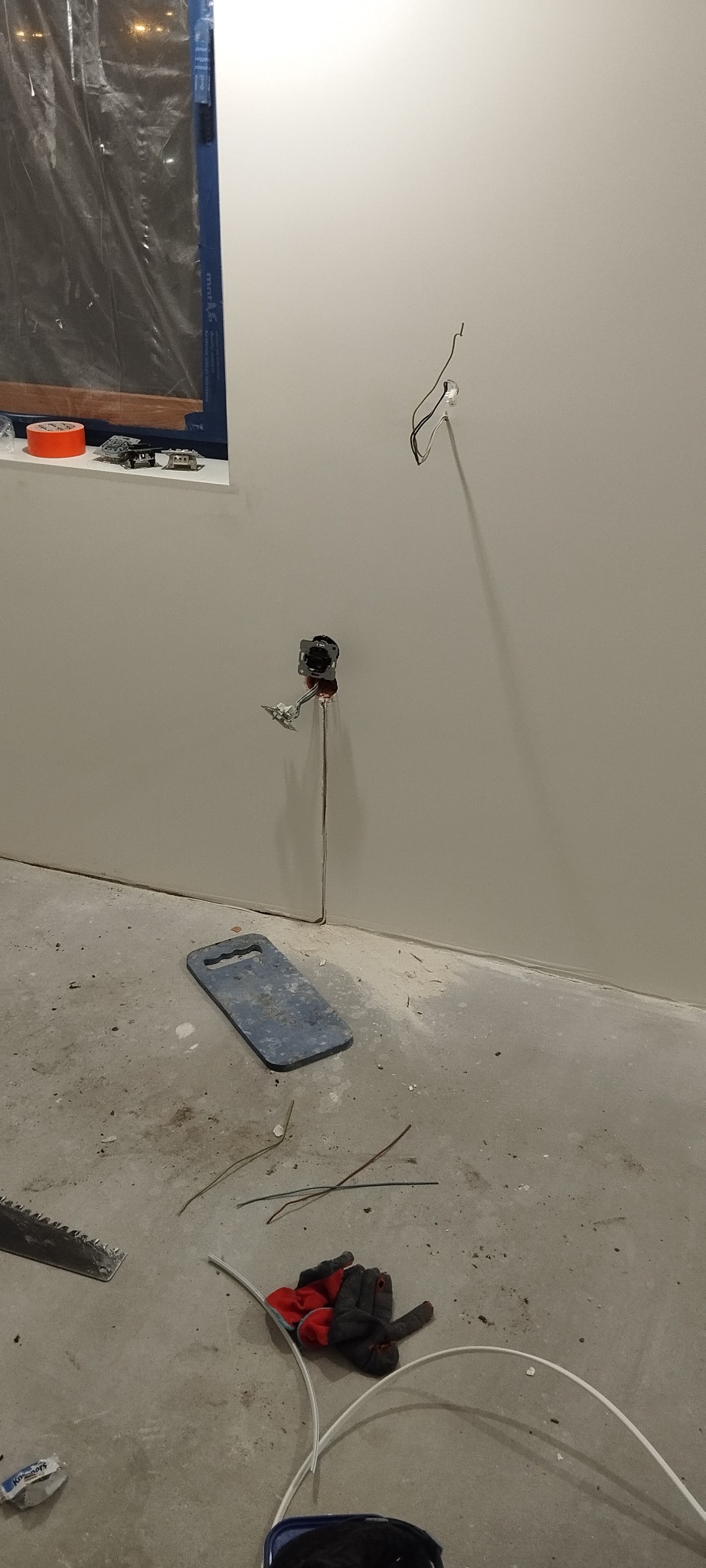 Przygotowanie do montażu gniazdka elektrycznego w nowym budynku, widoczne przewody wystające ze ściany, puszka instalacyjna oraz narzędzia i rękawice robocze na surowej podłodze.