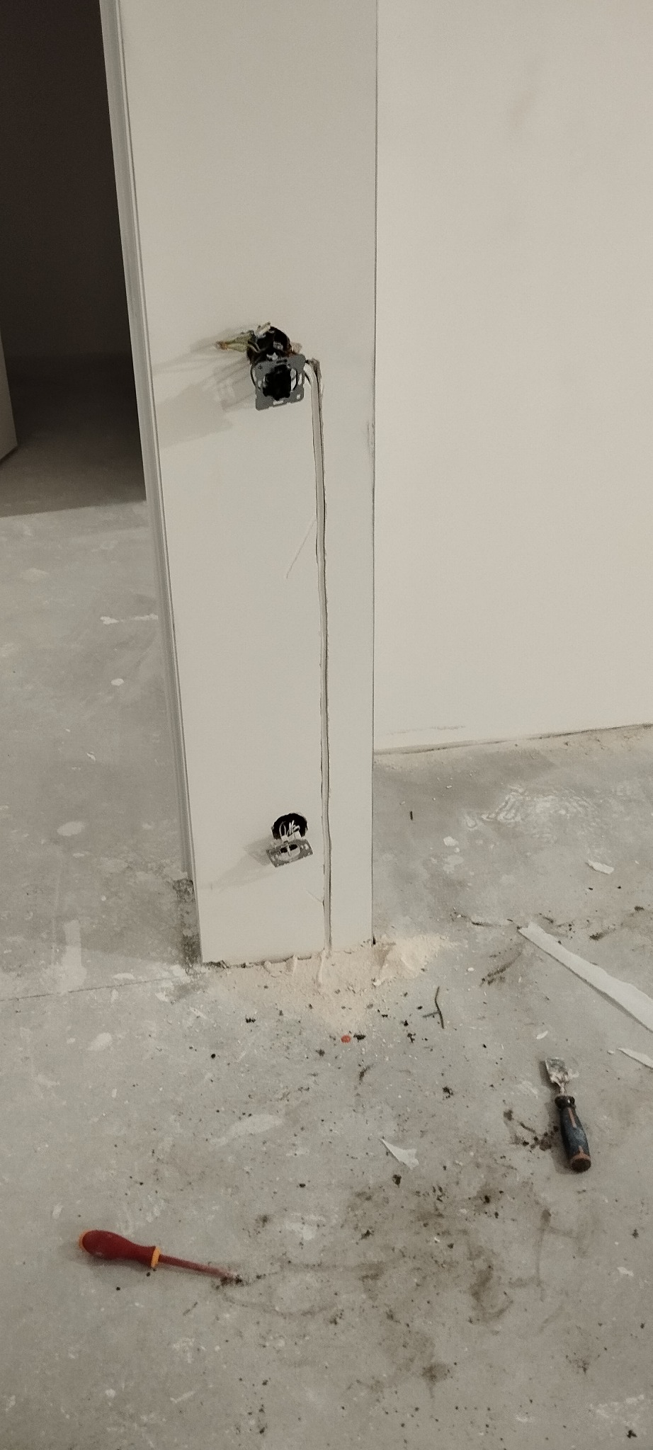 Przygotowanie do montażu gniazdek elektrycznych w ścianie, widoczne kable i puszki instalacyjne, na podłodze leżą narzędzia: śrubokręt i szpachelka.