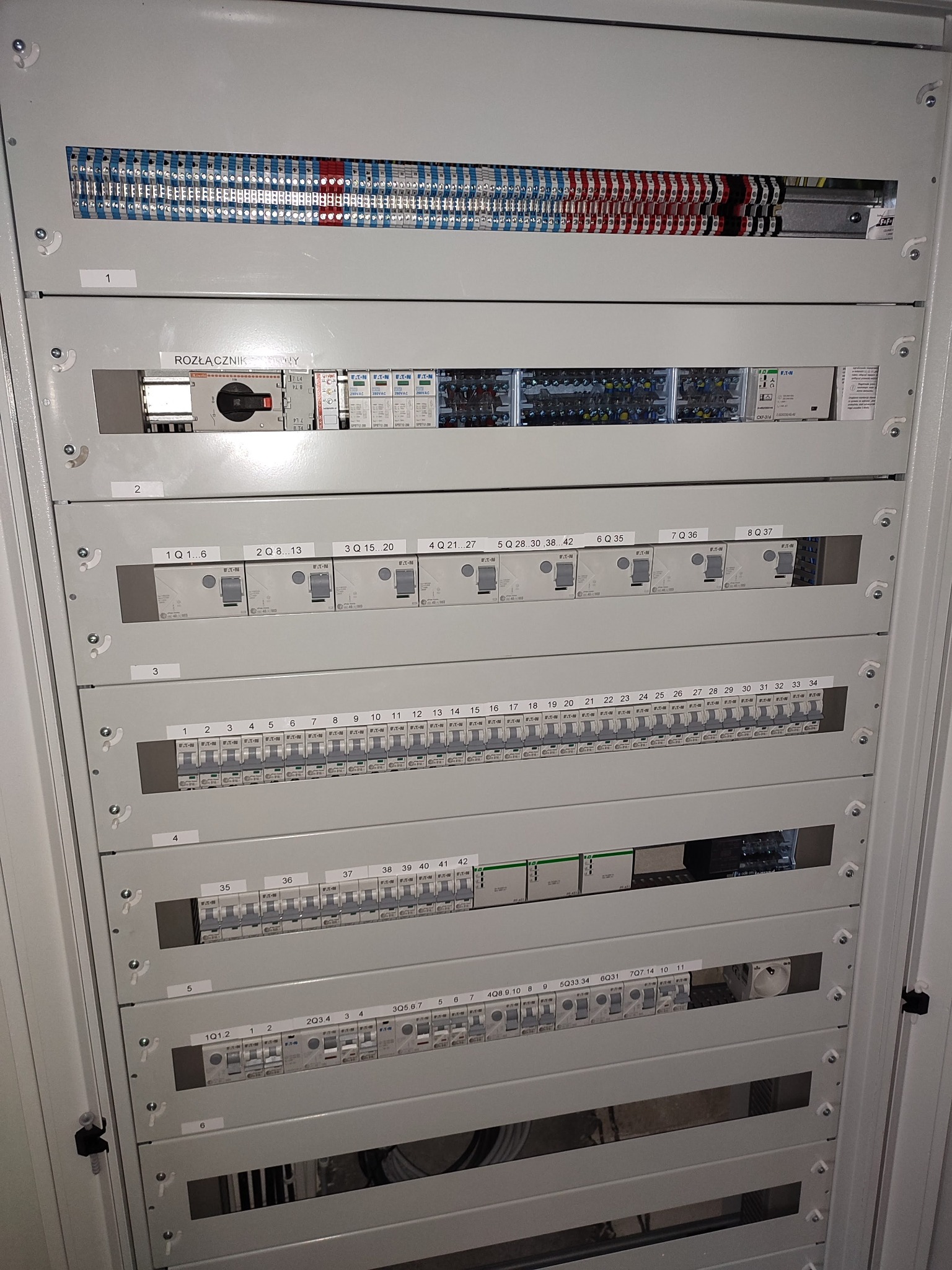 Rozdzielnica elektryczna z widocznymi bezpiecznikami, listwami zaciskowymi i wyłącznikiem, oznaczona numerami sekcji od 1 do 6, z opisami zakresów numerów bezpieczników w każdej sekcji.