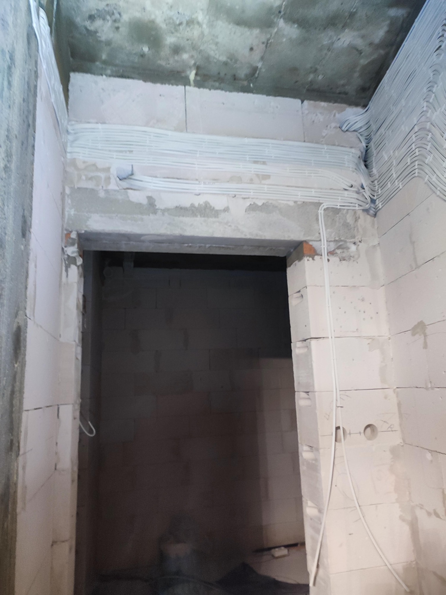 Surowy stan wnętrza budynku w trakcie prac elektrycznych: białe kable ułożone wzdłuż górnej części ściany z betonu komórkowego nad otworem drzwiowym, widoczne ślady po zaprawie i niedokończone...