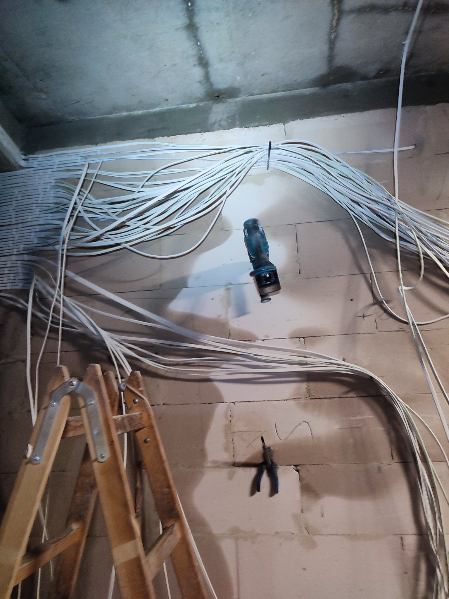 Rozprowadzenie kabli elektrycznych po ścianach i suficie surowego pomieszczenia, widoczna drabina, wiertarka i kombinerki w ścianie.