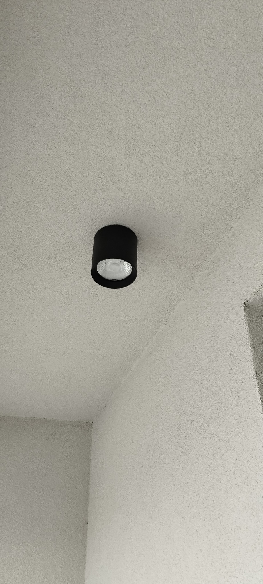 Czarna, okrągła lampa sufitowa typu downlight zamontowana na białym, chropowatym suficie w rogu pomieszczenia.