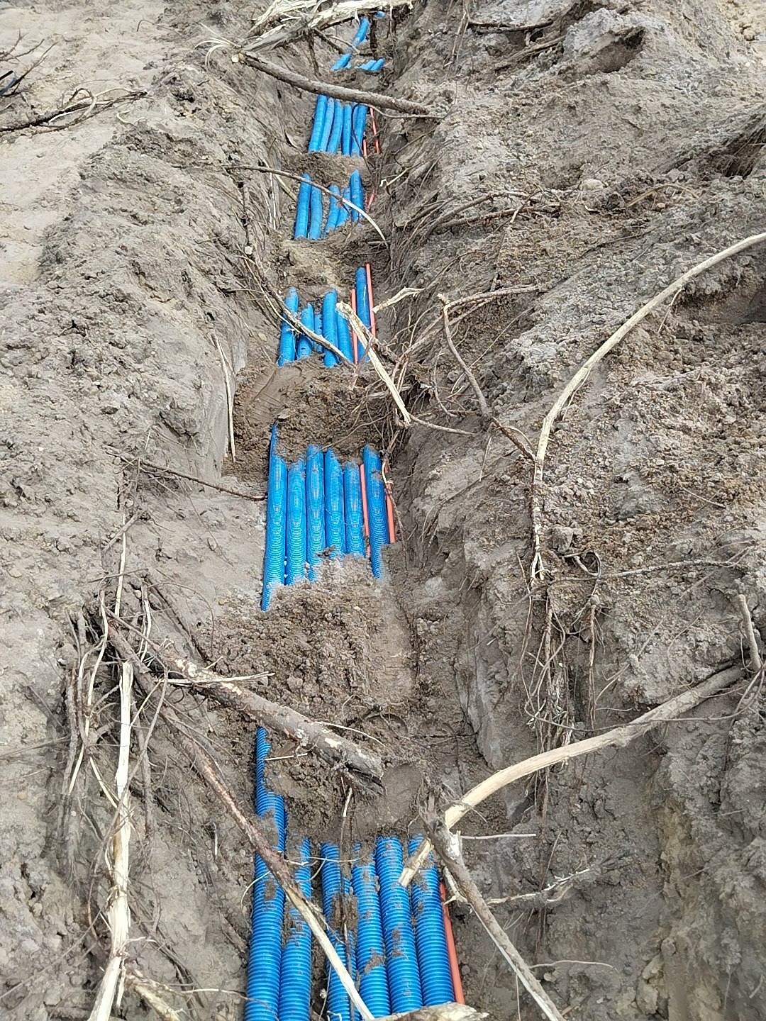 Niebieskie rury karbowane ułożone w wykopie ziemnym, częściowo przysypane, widoczne pomarańczowe kable. Instalacja podziemna przed zasypaniem.